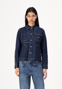 Veste en denim bleu foncé avec un design cropped, fermeture à boutons, poche poitrine et coutures contrastantes. Manches longues et col standard.