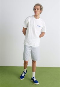 White cotton t-shirt with blue logo, light gray shorts, white socks, and blue sneakers. Standing on green carpet against a plain backdrop.