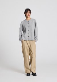 Cardigan grigio con bottoni, con orlo a coste e scollatura rotonda, abbinato a pantaloni beige a vestibilità ampia e scarpe nere, su uno sfondo semplice.