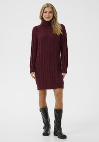 Burgundska pletena obleka s visokim ovratnikom, z značilnim kabelskim vzorcem in dolgimi rokavi. V kombinaciji z črnimi usnjenimi škornji do kolen.