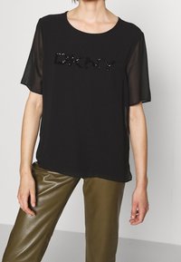 Top corto nero a maniche corte con scollatura rotonda e dettaglio in paillettes "DKNY". Tessuto morbido con maniche trasparenti, abbinato a pantaloni verde oliva.
