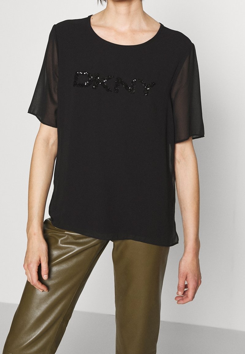 Top corto nero a maniche corte con scollatura rotonda e dettaglio in paillettes "DKNY". Tessuto morbido con maniche trasparenti, abbinato a pantaloni verde oliva.