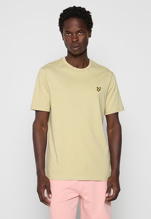 Homme portant un t-shirt jaune clair à manches courtes avec un petit logo d'oiseau jaune sur la poitrine et un pantalon rose clair, debout devant un fond uni.