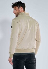 Beige Herren-Bomberjacke mit hohem Kragen und elastischen Bündchen. Verfügt über einen Aufnäher am linken Ärmel und einen gerippten Saum. Glatte Textur, leichtes Material.