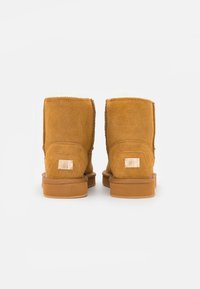 Bottines en daim beige avec une texture douce, présentant des coutures le long des coutures et un détail d'accent métallique à l'arrière.