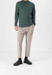 Grön långärmad polotröja, beige kordflossenbyxor med texturerad finish, och svarta kraftiga loafers, stående framför en enfärgad bakgrund.