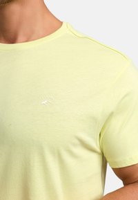 Kurzärmliges, hellgelbes T-Shirt aus weichem Stoff, mit einem runden Halsausschnitt und einem kleinen weißen Logo, das "EST. 1993." sagt.