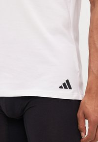 Weißes Baumwoll-T-Shirt mit geradem Schnitt, mit einem schwarzen Adidas-Logo nahe dem Saum, glatte Textur und kurzen Ärmeln.