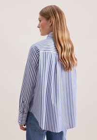Vrouw met lang blond haar, gekleed in een blauw-wit verticaal gestreept shirt en blauwe jeans, gezien van achteren en iets naar links gedraaid.