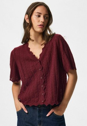 Femme portant un cardigan en tricot à manches courtes rouge foncé avec des bords festonnés et des boutons, associé à un jean bleu foncé, mains dans les poches.