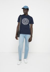 Marineblå t-shirt med cirkulært hvidt logo, lyseblå jeans, hvide sneakers og en lyseblå kasket. Model bærer en sort messenger taske.