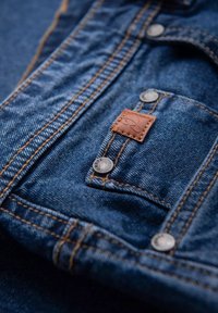 Läheltä kuvattua denimpintaa, jossa on kuparinsävyinen nahkakappale, hopeiset napit ja erottuva ommel syvällä sinisellä taustalla.