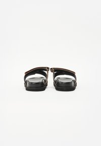 Sorte sandaler med brune teksturerte stropper, åpen tå-design, flate såler og minimale hardwaredetaljer.