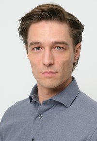 Camicia grigia con bottoni, realizzata in un tessuto testurizzato, con colletto classico e bottoni in vista. Il modello ha capelli di lunghezza media e arruffati.