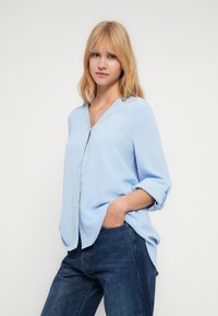 Ανοιχτό μπλε πουκάμισο με μακριά μανίκια και κουμπιά, V-λαιμόκοψη, υφασμάτινη υφή και γυρισμένα μανίκια, σε συνδυασμό με σκούρο τζιν παντελόνι.