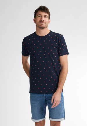 Marineblå t-shirt med rund hals og korte ærmer, prydet med et gentaget mønster af små røde designs. Båret sammen med denimshorts.