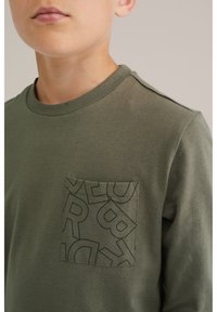 Camisa de manga larga verde de algodón con un bolsillo texturizado que presenta un patrón de letras repetido, escote acanalado y detalles en los hombros.