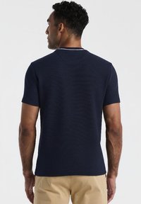 Homme aux cheveux courts et bouclés portant une chemise à manches courtes bleu marine et un pantalon beige, vu de dos sur un fond uni.