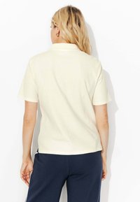 Polo à manches courtes de couleur crème clair avec une texture côtelée et un col classique, associé à un pantalon bleu foncé, vu de dos.