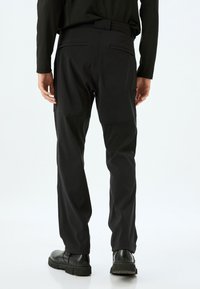 Sorte bukser med en glat tekstur, slim fit og forstærkede knæpaneler. Har lommer og bæltestropper, båret med sorte sko.