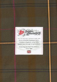 Brązowa tkanina w kratę z pomarańczowymi i purpurowymi paskami. Metka zawiera napis "The British Millerain Co Ltd" oraz szczegóły dotyczące jakości wykonania tkaniny.