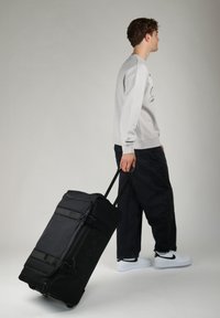 Borsa a duffel nera con ruote, maniglie superiori e laterali, dotata di chiusura con zip e tasca frontale, portata da una persona in abbigliamento casual.