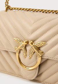 Μπεζ καπιτονέ δερμάτινη τσάντα με χρυσούς μεταλλικούς λεπτομέρειες που διαθέτει δύο πουλιά ως κούμπωμα. Περιλαμβάνει ένα στριφτό αλυσίδα ιμάντα.