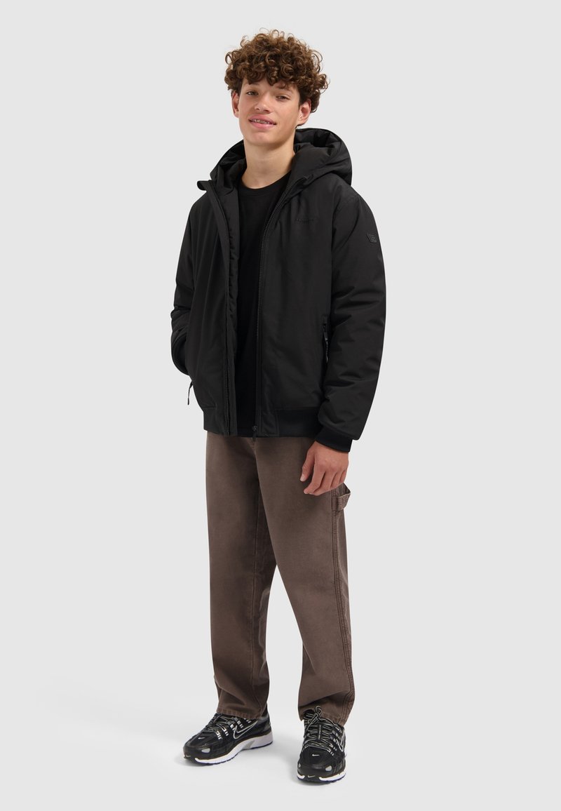 Tienerjongen staat in een zwarte hoodie, zwart shirt, bruine broek en zwarte sneakers tegen een effen lichte achtergrond.