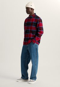 Rood en zwart geruit flanellen shirt met blauwe accenten, gecombineerd met lichtblauwe jeans. Witte canvas schoenen met een ruitpatroon, geribbelde witte beanie.
