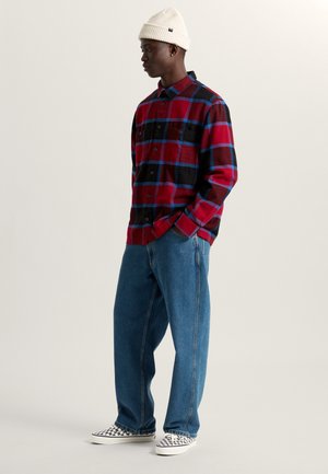 Camicia di flanella a quadri rossi e neri con accenti blu, abbinata a jeans azzurri chiari. Scarpe da ginnastica bianche con un motivo a scacchi, berretto bianco a costine.