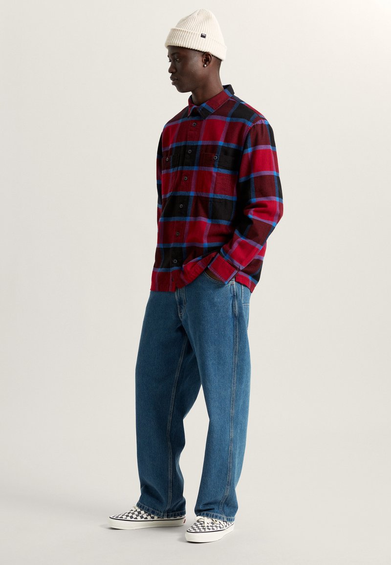 Rood en zwart geruit flanellen shirt met blauwe accenten, gecombineerd met lichtblauwe jeans. Witte canvas schoenen met een ruitpatroon, geribbelde witte beanie.