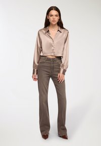 Blusa in satin color beige chiaro con colletto e chiusura a bottoni, abbinata a jeans svasati marrone scuro. Entrambi i capi presentano texture morbide e linee pulite.