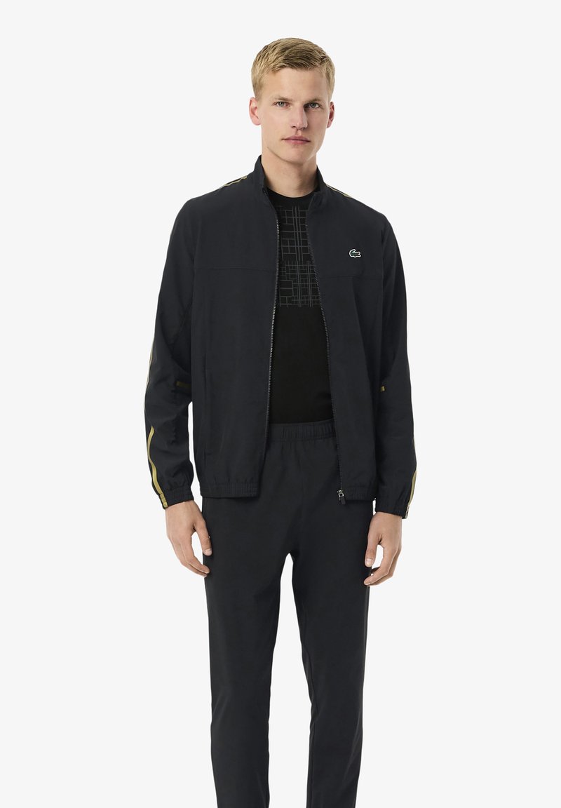 Junger Mann trägt eine schwarze Zip-Jacke mit goldenen Streifen an den Ärmeln, ein schwarzes bedrucktes T-Shirt und schwarze Hosen, steht vor einem weißen Hintergrund.