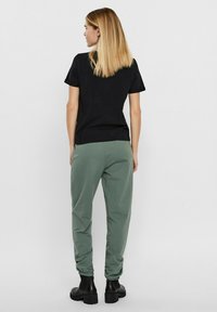 Schwarzes Baumwoll-T-Shirt mit kurzen Ärmeln, kombiniert mit grünen Jogginghosen. Die Hosen haben einen elastischen Bund, und das Model trägt schwarze Knöchelstiefel.