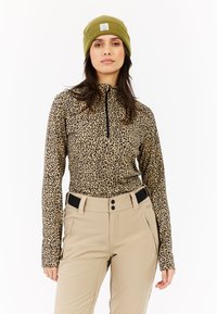 Camiseta de manga larga con estampado de leopardo y cremallera negra, combinada con pantalones beige con acentos negros y bolsillos delanteros. Gorro verde oliva en la cabeza.