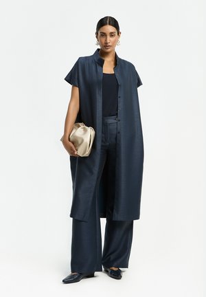 Frau trägt einen marineblauen, ärmellosen langen Mantel, passende weit geschnittene Hosen, ein schwarzes Oberteil und hält eine beige Clutch, steht vor weißem Hintergrund.