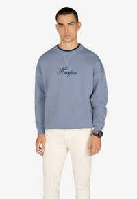 Felpa azzurra con polsi a costine, collo rotondo e ricamo "Harper" in blu navy. Indossata con jeans bianchi aderenti.