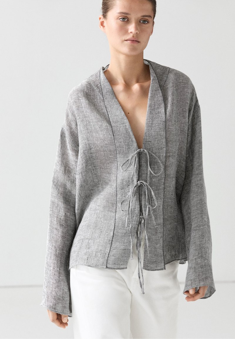 Frau trägt eine lockere graue Leinenstrickjacke mit zwei vorderen Bändern, kombiniert mit weißen Hosen, vor schlichten hellem Hintergrund.