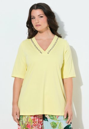 Vrouw met golvend donker haar, gekleed in een gele V-hals top met strass-steentjes en kleurrijke broek met bloemenpatroon, staand tegen een witte achtergrond.