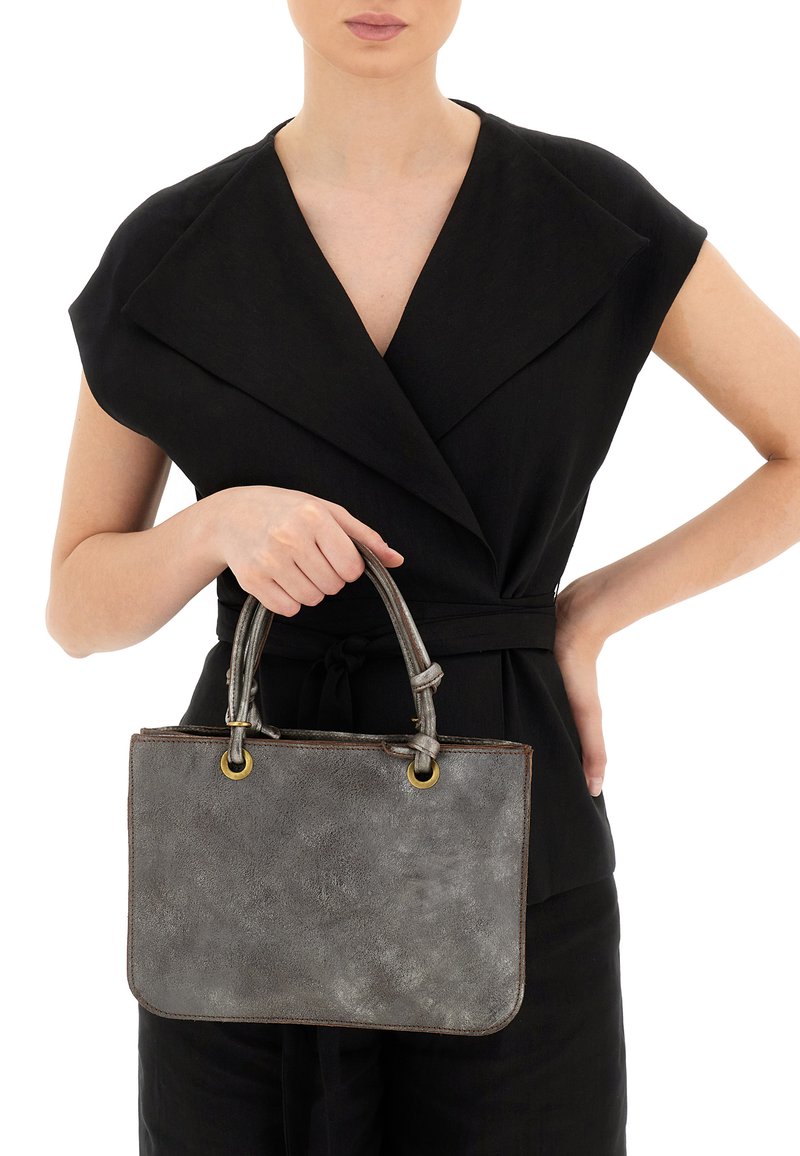 Borsa in pelle grigia con due manici superiori, caratterizzata da hardware dorato, una superficie liscia e angoli arrotondati. Il modello tiene la borsa davanti a sé.