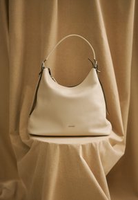 Bež usnjena hobo torba s strukturirano obliko, enim ozkim ročajem in srebrnimi kovinskimi poudarki. Teksturirana zunanjost in viden logotip.