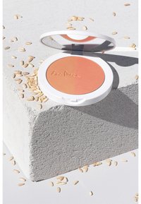 Compact blush in een witte doos, met een tweekleurig poeder in perzik- en koraaltinten. Reflecterende spiegel aan de binnenkant, omgeven door verspreide korrels.