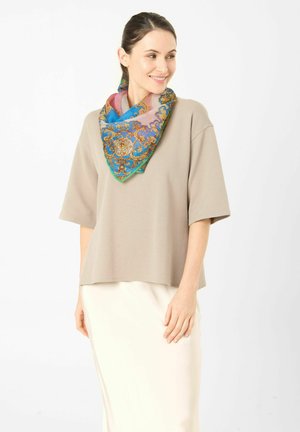 Vrouw draagt een beige losse top, witte rok en kleurrijke sjaal met patroon, glimlachend en kijkend naar de zijkant tegen een effen achtergrond.