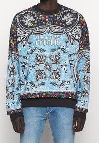 Blå och svart mönstrad sweatshirt med blommönster, paisleymotiv och en logotyp. Svarta ribbade muddar och midjeband fulländar designen.