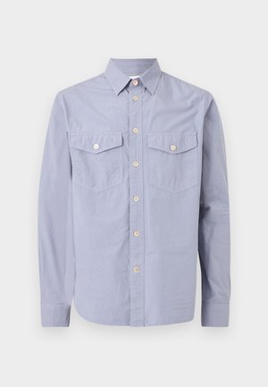 Junger Mann mit lockigem Haar, der ein hellblaues Hemd mit Knöpfen, ein weißes T-Shirt, dunkle, knielange Jeansshorts, weiße Socken und weiße Sneaker trägt.