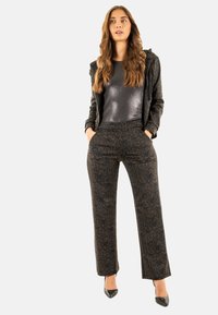 Pantalones de estampado de leopardo en negro y marrón, combinados con una blusa de textura negra y una chaqueta con cremallera a juego. La modelo lleva tacones negros.