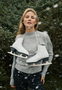 Baltas ledus skrituļslidas karājas uz auklas ap cilvēka kaklu, kurš valkā pelēku džemperi ar rotājumiem. Fonā krīt sniegs.