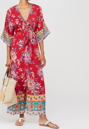 Robe rouge à fleurs avec un devant noué, des manches courtes larges et une bordure colorée à motifs. Associée à un sac fourre-tout tressé et des sandales plates.