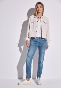 Veste courte beige clair avec deux poches poitrine, associée à un jean bleu clair déchiré avec des patches. Le modèle porte des baskets blanches.