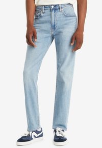 Uomo che indossa jeans dritti azzurri e sneakers blu navy con lacci bianchi, in piedi su uno sfondo bianco.